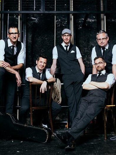A band with five members poses in elegant clothing. In the background, musical instruments and stage lighting can be seen. | © Bild: Marija Kanizaj