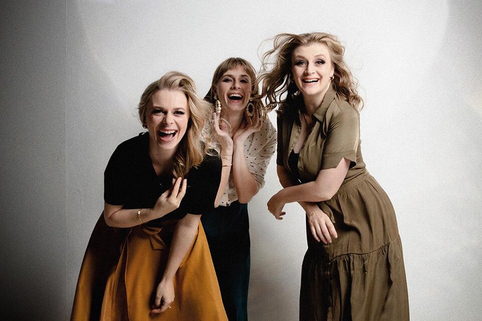 Drei Frauen lächeln fröhlich und posieren vor einem neutralen Hintergrund. Sie tragen verschiedene Kleidung und scheinen viel Spaß zu haben. | ©  Zoe Goldstein Fotografie