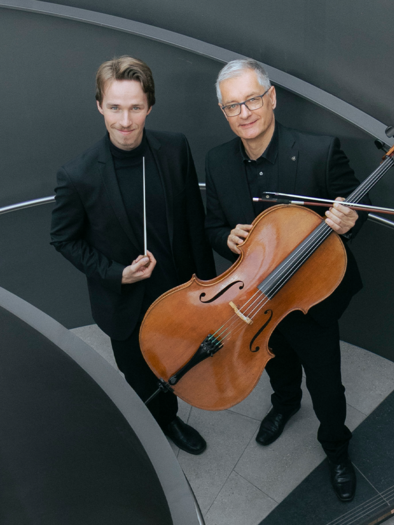 Two musicians are standing on a staircase and posing with a cello. The man on the left is holding a conductor's baton. | ©  leopress