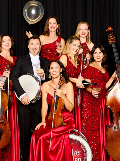 A group of musicians in glittering red dresses stands together. They hold instruments and appear festive and elegant. | ©  Kurt Remling