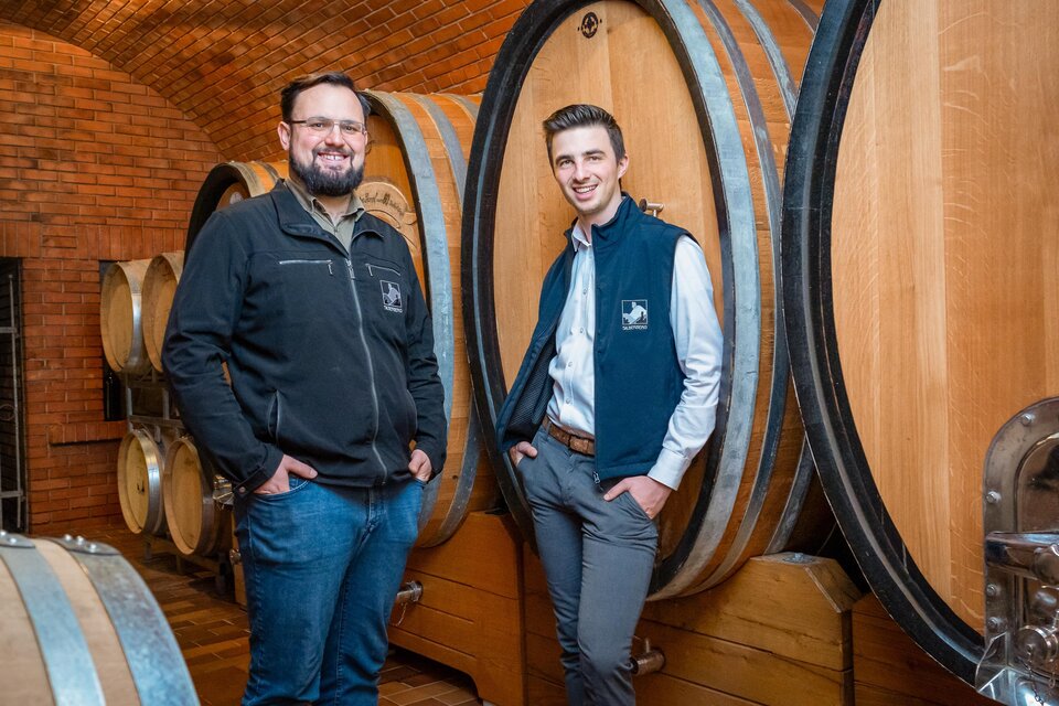 Zwei Männer stehen in einem Weinkeller neben Holzfässern. Die Atmosphäre ist rustikal und einladend. | © Silberberg_Oenologe Menhart_Kellermeister Lobe_Fasskeller_Mario Gimpel