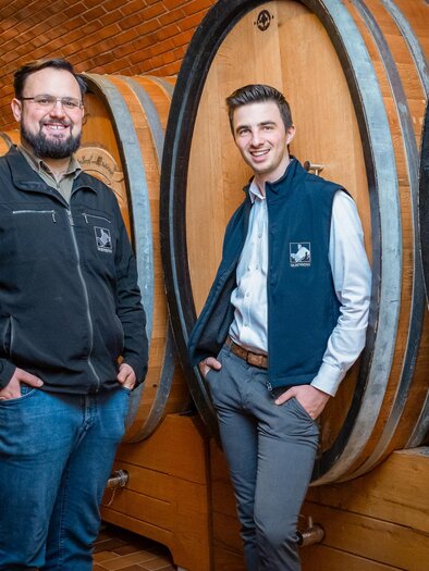 Two men are standing in a wine cellar next to wooden barrels. The atmosphere is rustic and inviting. | © Silberberg_Oenologe Menhart_Kellermeister Lobe_Fasskeller_Mario Gimpel
