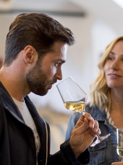 A man and a woman are having a conversation in a wine shop. The man is sniffing a wine glass while the woman watches with interest. | © TV Südsteiermark