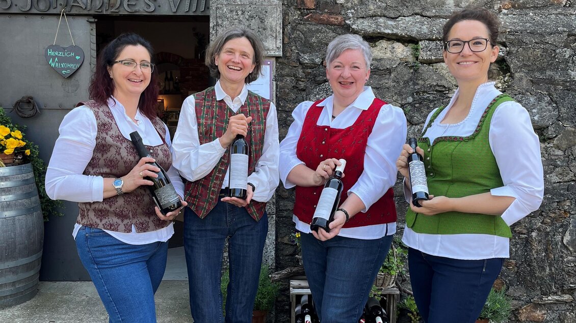 Vier Frauen stehen vor einem historischen Gebäude und halten Weinflaschen in der Hand. Sie tragen traditionelle Trachten und lächeln freundlich in die Kamera.