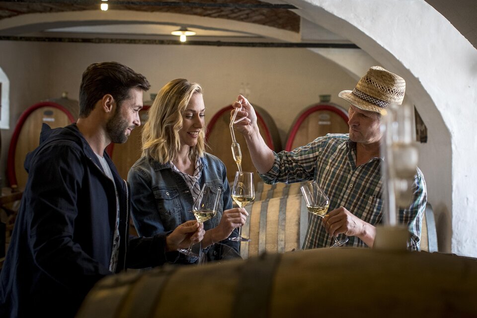 Drei Personen stehen in einem Weinkeller und probieren Wein. Der Winzer schenkt ihnen mit einem Löffel Wein ein. | © Tom Lamm