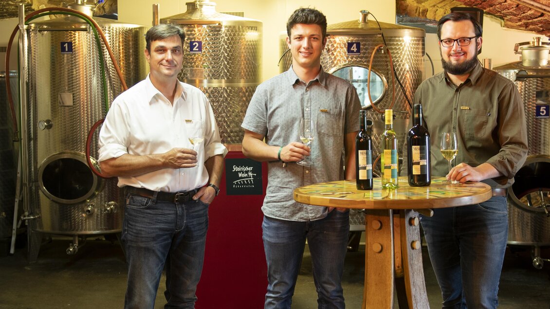 Drei Männer stehen an einem Tisch mit Weinflaschen in einem Weinkeller. Im Hintergrund sind große Edelstahltanks zu sehen.