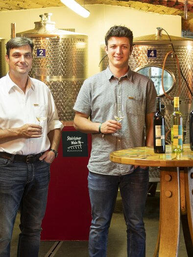 Three men are standing at a table with wine bottles in a wine cellar. In the background, large stainless steel tanks can be seen.