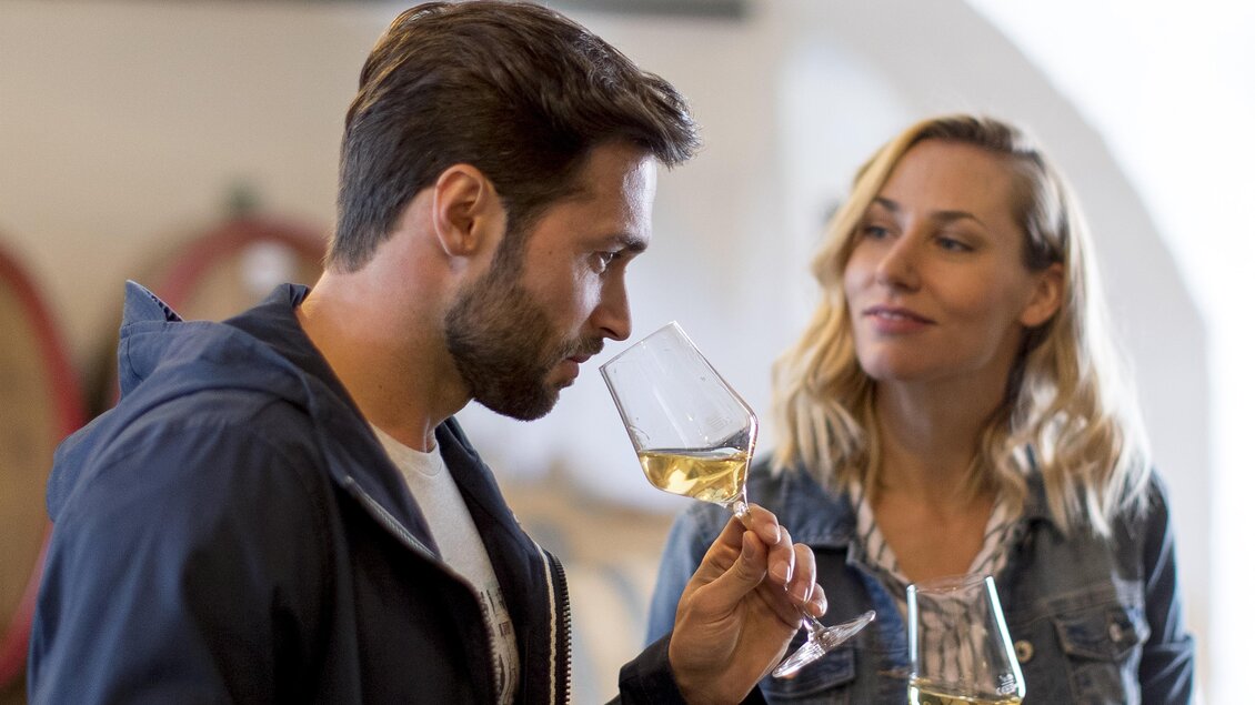 Ein Mann und eine Frau unterhalten sich in einem Weingeschäft. Der Mann schnuppert an einem Weinglas, während die Frau interessiert zuschaut. | © TV Südsteiermark