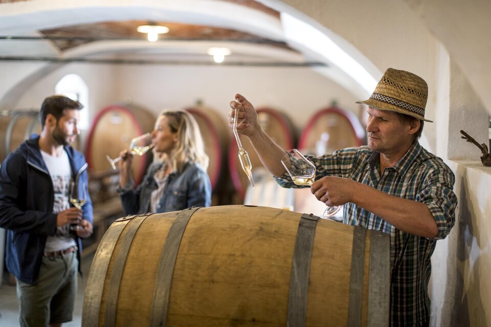 Eine Weinprobe in einem Weinkeller mit einem Winzer, der Wein aus einem Fass einschenkt. Im Hintergrund stehen zwei Personen, die ein Glas Wein genießen. | © Tom Lamm