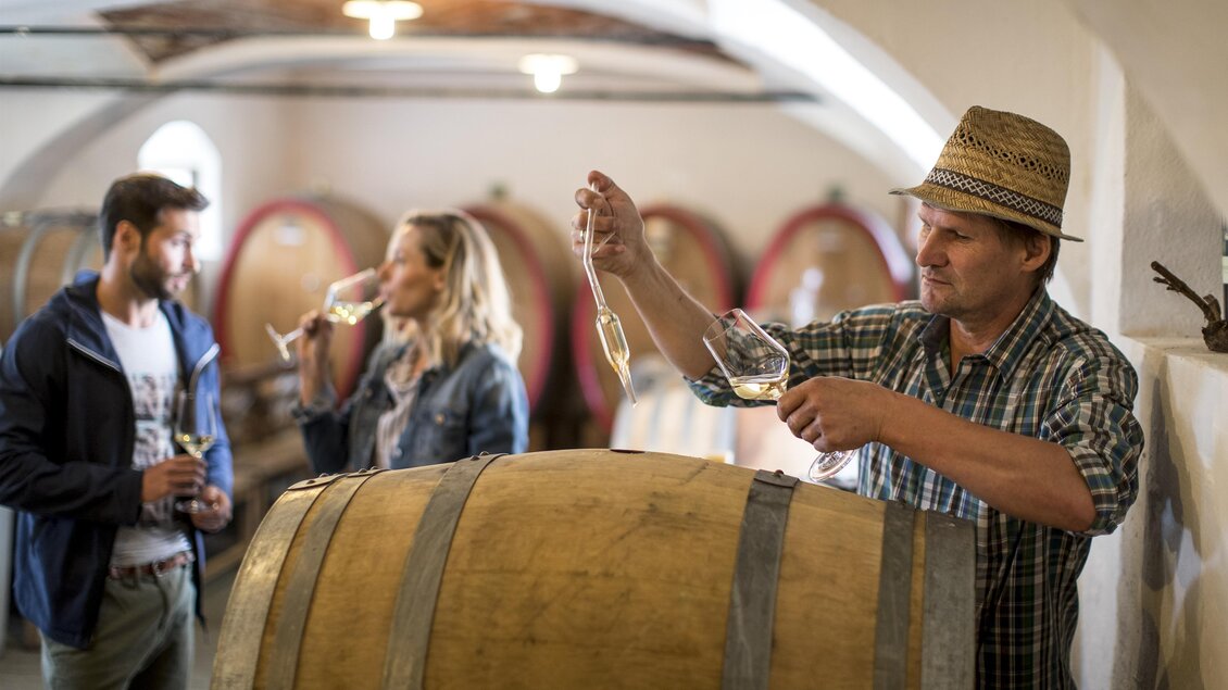 Eine Weinprobe in einem Weinkeller mit einem Winzer, der Wein aus einem Fass einschenkt. Im Hintergrund stehen zwei Personen, die ein Glas Wein genießen. | © Tom Lamm