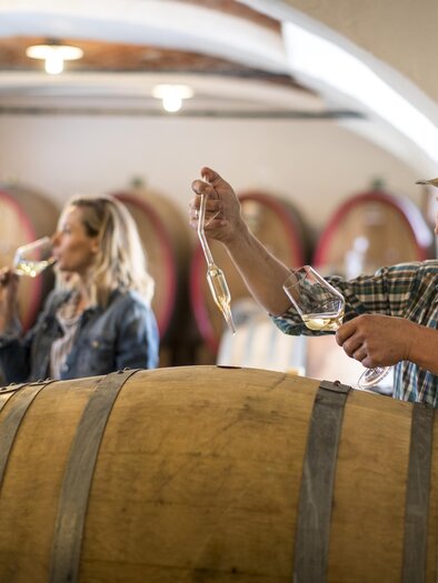 A wine tasting in a wine cellar with a winemaker pouring wine from a barrel. In the background, two people are enjoying a glass of wine. | © Tom Lamm