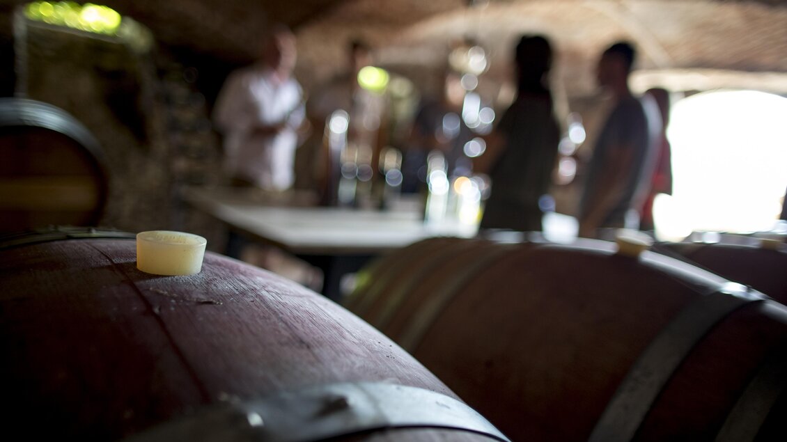 Ein Weinkeller mit Holzfässern im Vordergrund. Im Hintergrund sind Menschen, die sich unterhalten und Wein verkosten. | © Tom Lamm