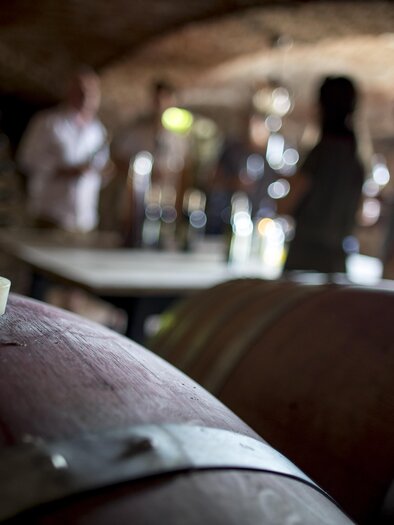 A wine cellar with wooden barrels in the foreground. In the background, there are people talking and tasting wine. | © Tom Lamm