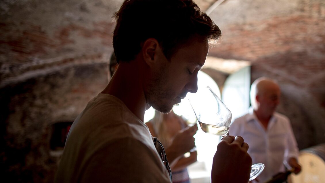 Ein Mann riecht an einem Weinglas in einem Weinkeller. Im Hintergrund sind weitere Personen und Weinfässer sichtbar. | © Tom Lamm