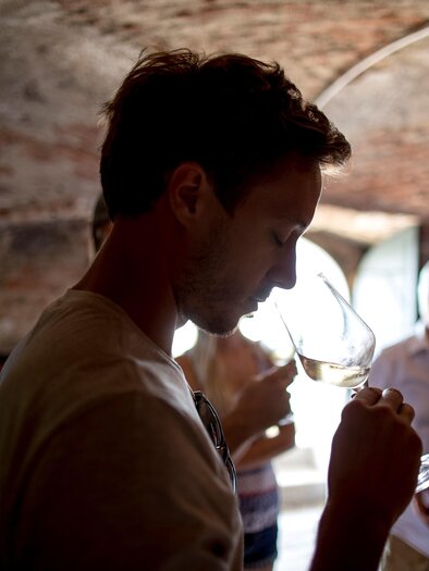 A man is smelling a wine glass in a wine cellar. In the background, other people and wine barrels are visible. | © Tom Lamm