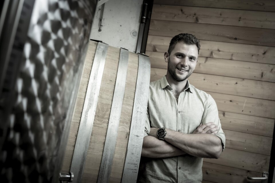 Ein Mann steht lächelnd neben einem Holzfass in einem Raum mit Holzwänden. Er trägt ein Hemd und hat die Arme verschränkt. | © Fam. Narat-Zitz