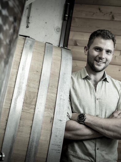 Ein Mann steht lächelnd neben einem Holzfass in einem Raum mit Holzwänden. Er trägt ein Hemd und hat die Arme verschränkt. | © Fam. Narat-Zitz