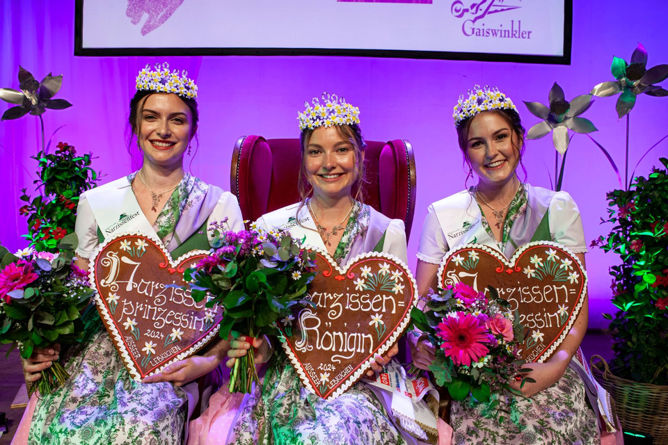 Drei Frauen in traditioneller Tracht sitzen auf einem roten Thron. Jede hält ein herzförmiges Schild und hat einen Blumenstrauß in der Hand. | © www.narzissenfest.at-Sima