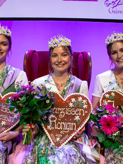 Three women in traditional attire are sitting on a red throne. Each holds a heart-shaped shield and has a bouquet of flowers in their hand. | © www.narzissenfest.at-Sima
