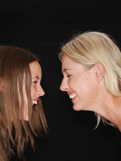 A laughing woman and a girl are facing each other. Both are joyful and seem to be sharing a fun moment.