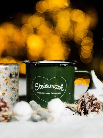 A green cup with the inscription "Steiermark" stands in the snow, surrounded by a glow of light and decorative elements such as pine cones. In the background, there are blurred lights that create a festive atmosphere. | © Schilcherland Steiermark