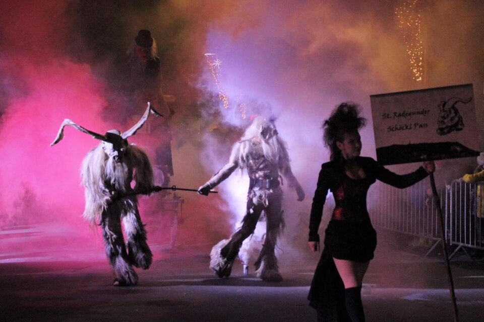 Eine bunte Parade mit mystischen Kreaturen und einer Frau, die ein Schild hält. Bunte Rauchschwaden und nächtliche Beleuchtung schaffen eine geheimnisvolle Atmosphäre. | © St. Radegunder Schöcklpass