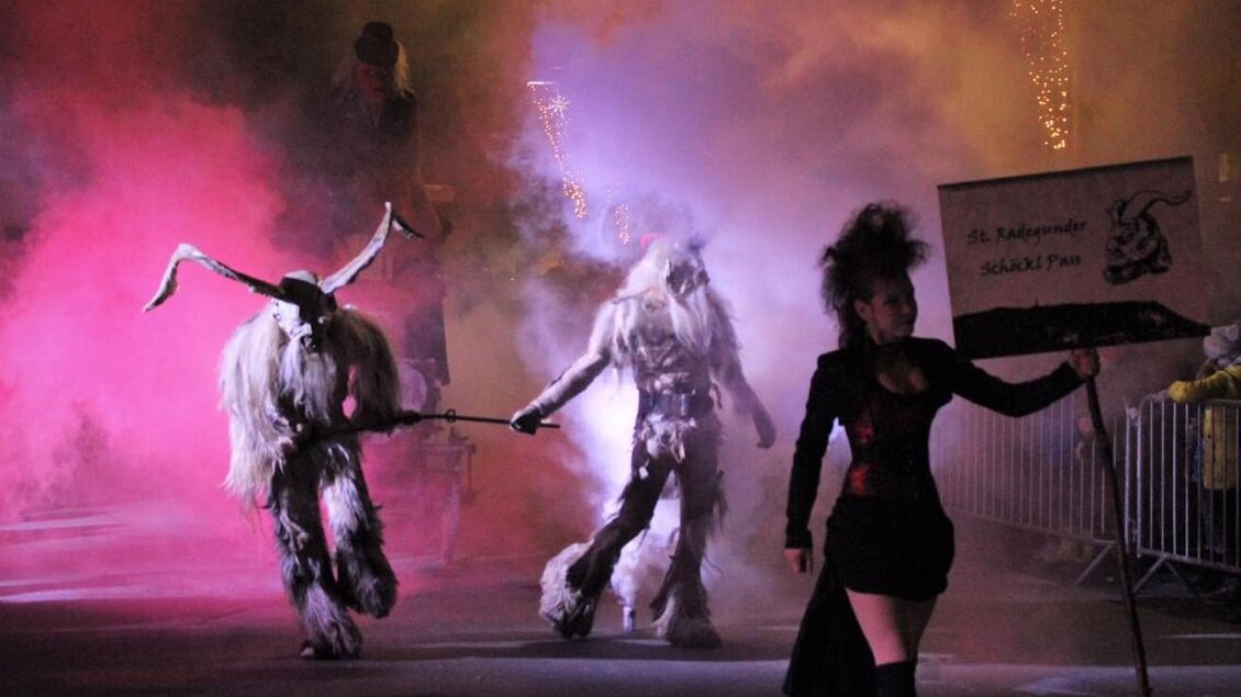 Eine bunte Parade mit mystischen Kreaturen und einer Frau, die ein Schild hält. Bunte Rauchschwaden und nächtliche Beleuchtung schaffen eine geheimnisvolle Atmosphäre. | © St. Radegunder Schöcklpass
