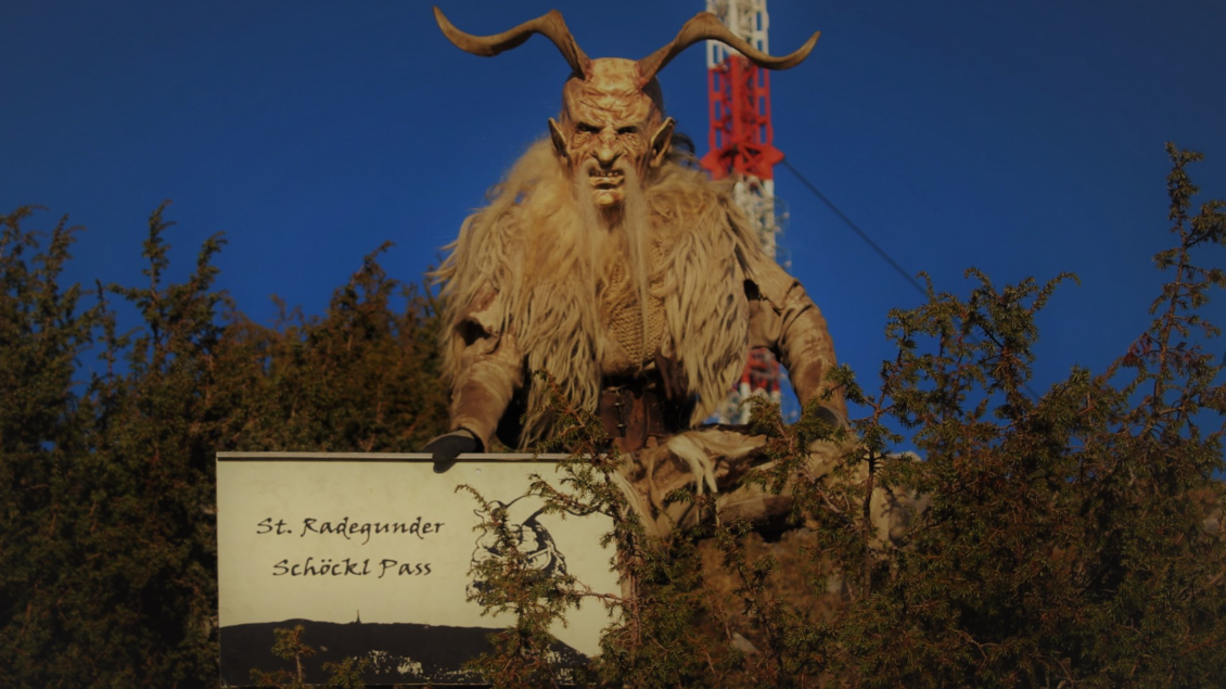 Eine beeindruckende Statue eines mystischen Wesens mit Hörnern, die vor einem Schild steht. Der Himmel ist klar und es sind einige Bäume im Hintergrund sichtbar. | © @ St. Radegunder Schöcklpass