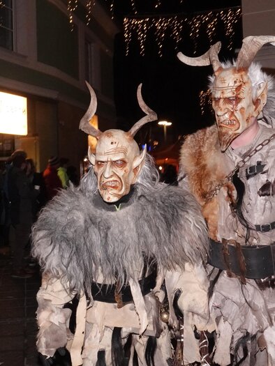 Two people in imaginative costumes with horns and frightening masks are standing on a street. In the background, other spectators can be seen observing the scene. | © Tourismusverband Oststeiermark