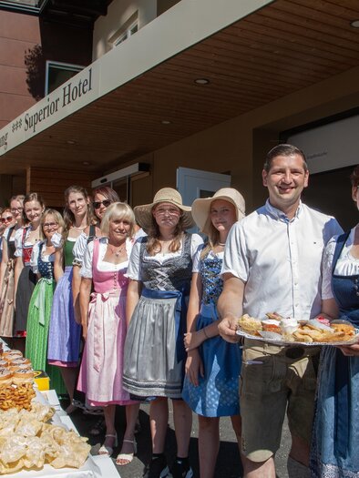A group of people in traditional costumes stands in front of a festively laid table with various baked goods. The atmosphere is friendly and inviting. | © Tourismusverein Mönichwald
