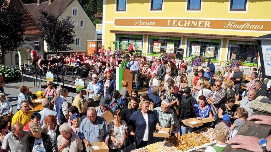 Eine lebhafte Menschenmenge versammelt sich vor einem Geschäft, um verschiedene Leckereien zu probieren. Die Straße ist festlich geschmückt und führt zu einer bunten Atmosphäre. | © Heinz Bayer