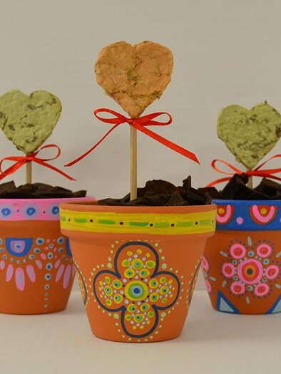Three colorful flower pots with decorative hearts on sticks are standing on a table. Each pot is uniquely painted and filled with soil. | ©  KulturQuartier Leoben