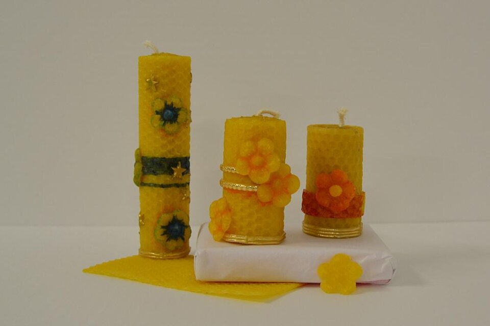 Drei dekorative gelbe Kerzen mit Blumenmotiven stehen auf einem weißen Untergrund. Neben den Kerzen liegt ein gelbes Blatt Wachs. | © KulturQuartier Leoben