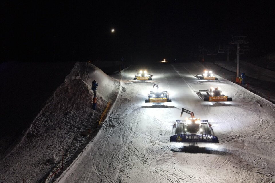 Mehrere Pistenraupen arbeiten nachts auf einer verschneiten Skipiste. Die Maschinen sind mit Scheinwerfern beleuchtet und formen die Schneedecke.