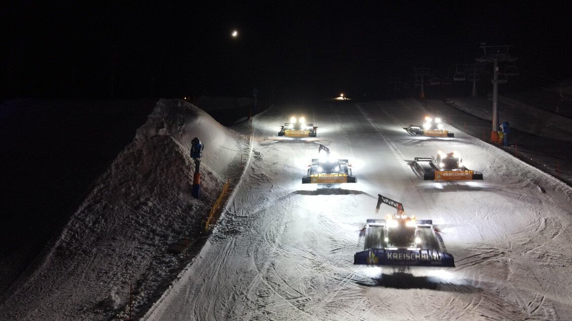 Mehrere Pistenraupen arbeiten nachts auf einer verschneiten Skipiste. Die Maschinen sind mit Scheinwerfern beleuchtet und formen die Schneedecke.