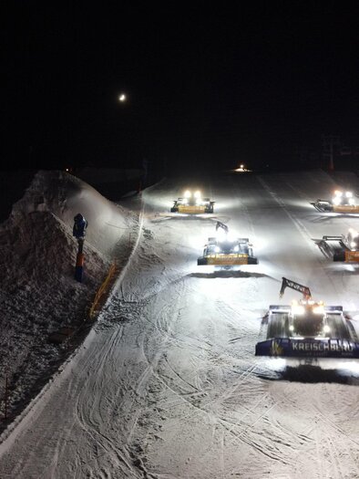 Several snow groomers work at night on a snow-covered ski slope. The machines are illuminated with headlights and shape the snow surface.
