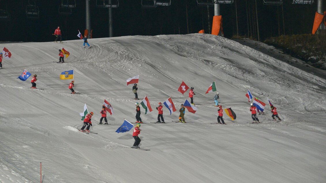 Eine Gruppe von Skifahrern fährt eine Piste hinunter und trägt verschiedene Flaggen. Im Hintergrund sind Skilifte sichtbar.