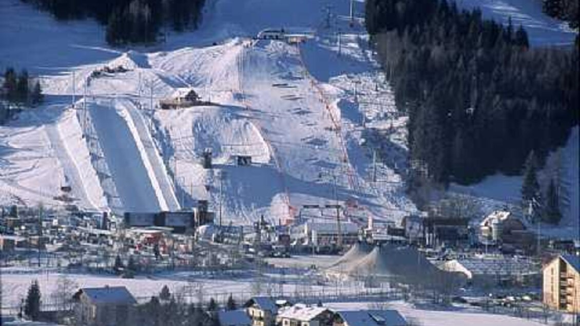 Eine schneebedeckte Landschaft mit mehreren Skipisten und einer Talstation. Im Hintergrund befinden sich Bäume und kleine Gebäude.