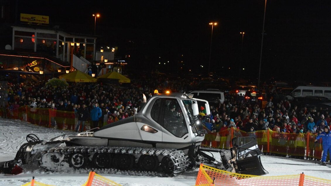 Ein Schneemobil steht auf einer verschneiten Fläche, umgeben von einer großen Menschenmenge bei Nacht. Im Hintergrund sind Lichter und Gebäude zu sehen.