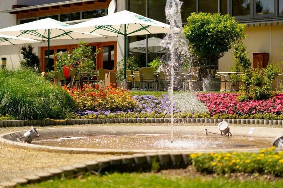 Springbrunnen im blühenden Garten vor einer Terrasse mit Sitzplätzen und Sonnenschirmen an einem schönen Tag. | © © Riedenbauer TVB Bad Waltersdorf