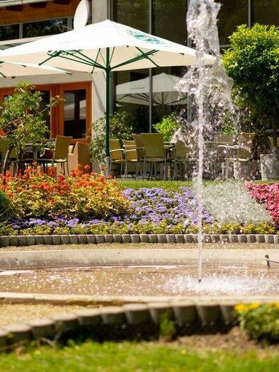 Fountain in a blooming garden in front of a terrace with seating and sun umbrellas on a beautiful day. | © © Riedenbauer TVB Bad Waltersdorf