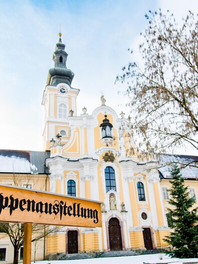 An impressive church with a tall tower and beautiful architecture. In front of the church stands a sign with the inscription "Nativity Scene Exhibition." | © TV Region Graz - Mias Photoart