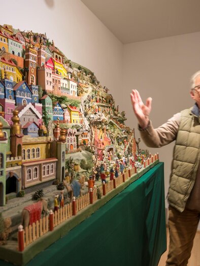 Ein älterer Mann steht neben einer bunten Miniaturstadt mit vielen Häusern und Figuren. Die Szenerie ist künstlerisch gestaltet und wirkt lebendig. | © Tourismusverband Oststeiermark