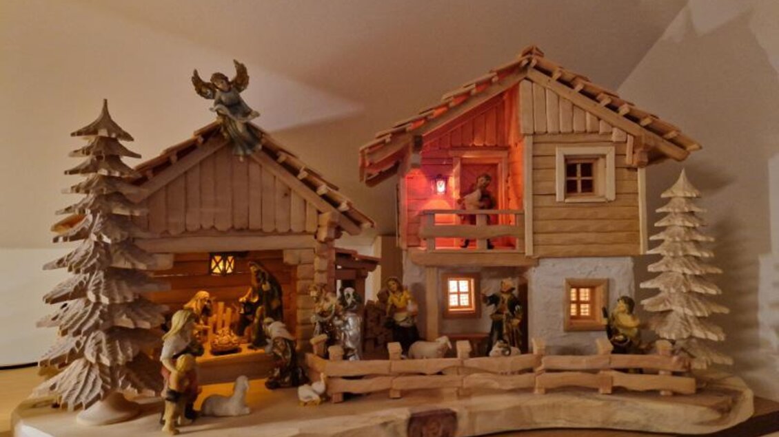 Eine festliche Weihnachtskrippe mit zwei Holzhäusern und Figuren. Das Szenario ist in warmem Licht beleuchtet und mit Tannenbäumen geschmückt. | © Adi Barazutti