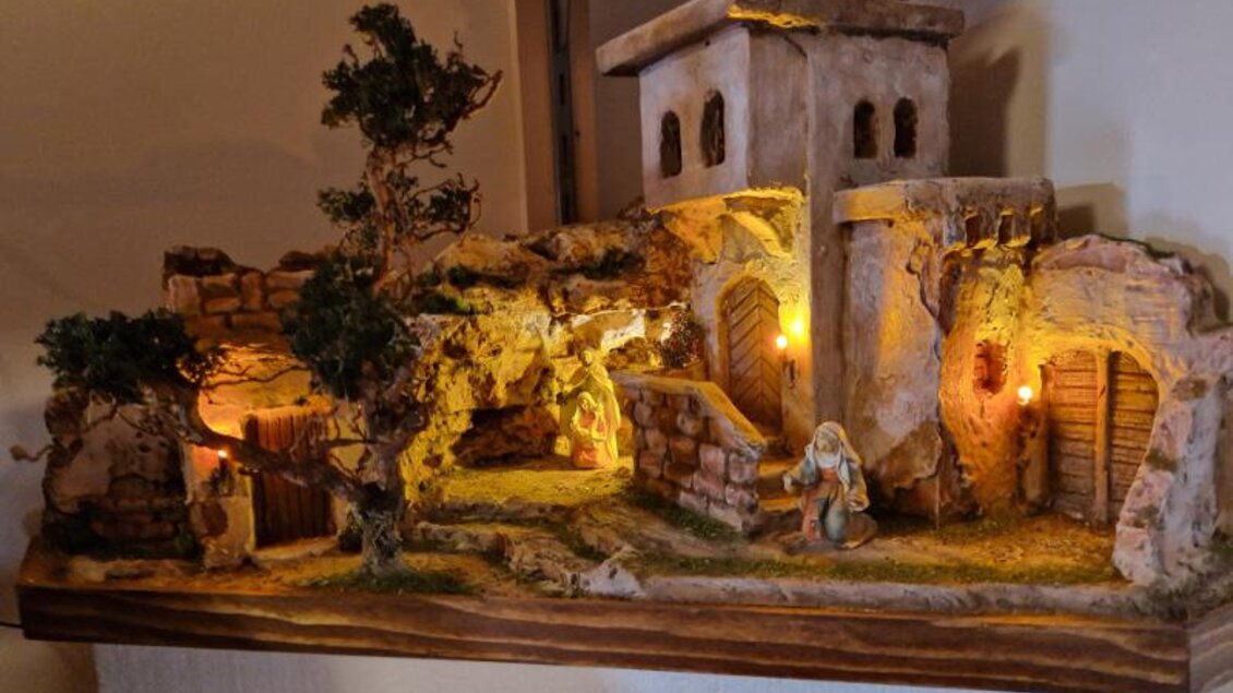 Eine dekorative Krippe mit beleuchteten Stallungen und einer Figur in einer natürlichen Umgebung. Der Hintergrund zeigt eine alte, steinige Architektur mit Bäumen. | © Adi Barazutti