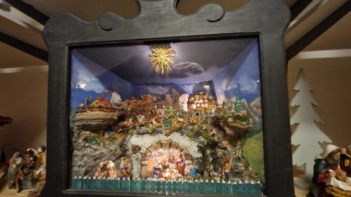 Eine kunstvoll gestaltete Krippe in einem großen schwarzen Rahmen. Die Szene zeigt ein idyllisches Dorf mit Bergen, Figuren und einer leuchtenden Sonne. | © Adi Barazutti