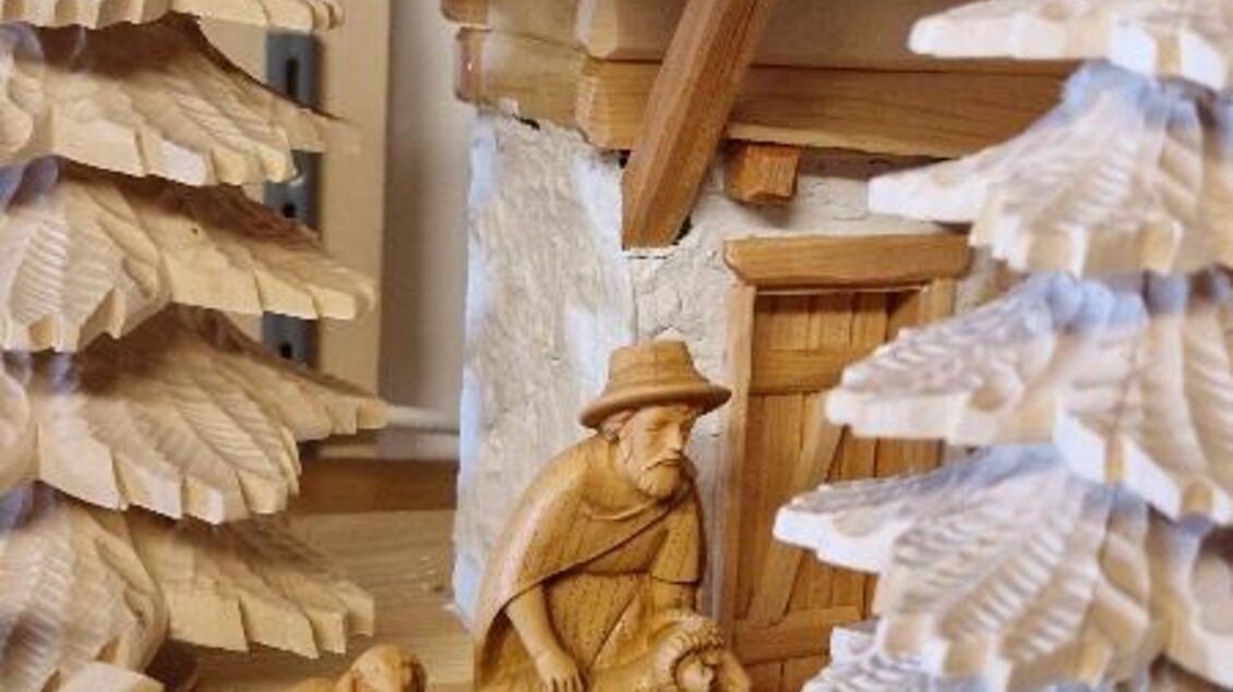 Eine kunstvoll geschnitzte Holzfigur eines Hirten mit Schafen vor einem kleinen Holzhaus, umgeben von Tannenbäumen. Die Details der Schnitzerei sind fein ausgearbeitet und zeigen ländliche Idylle. | © Adi Barazutti