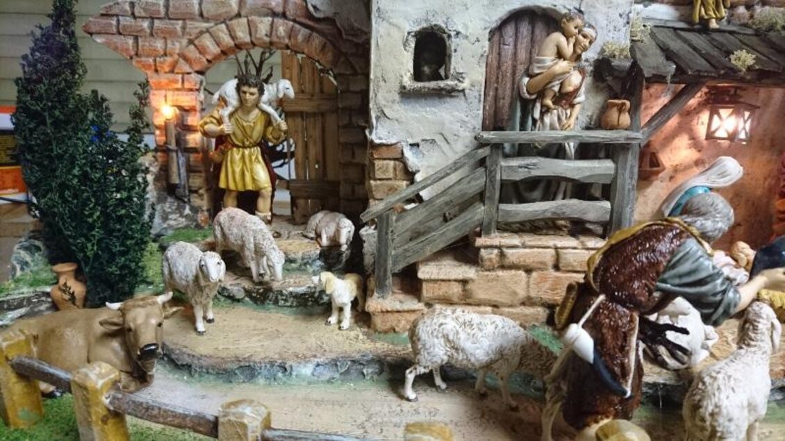 Eine traditionelle Krippenszene mit Hirten, Schafen und einem Stall. Im Hintergrund steht eine Figur mit einem Kind und dekorative Elemente. | © Adi Barazutti