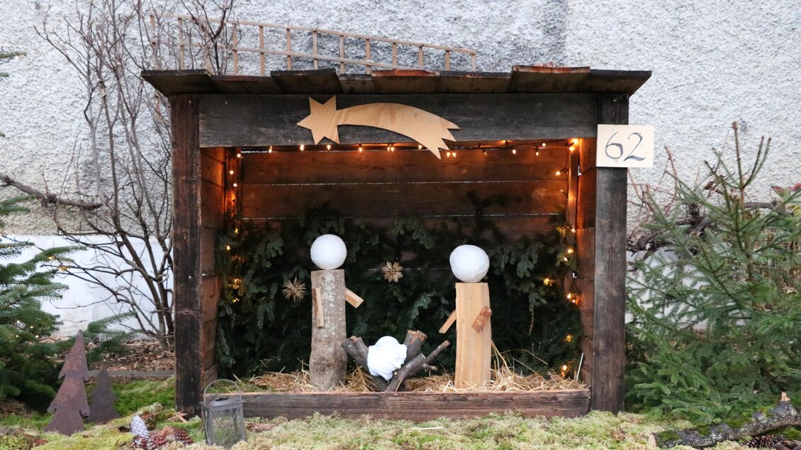 Eine festliche Weihnachtskrippe mit Holzfiguren und Lichtern. Im Hintergrund sind Tannenzweige zu sehen, die eine gemütliche Atmosphäre schaffen.