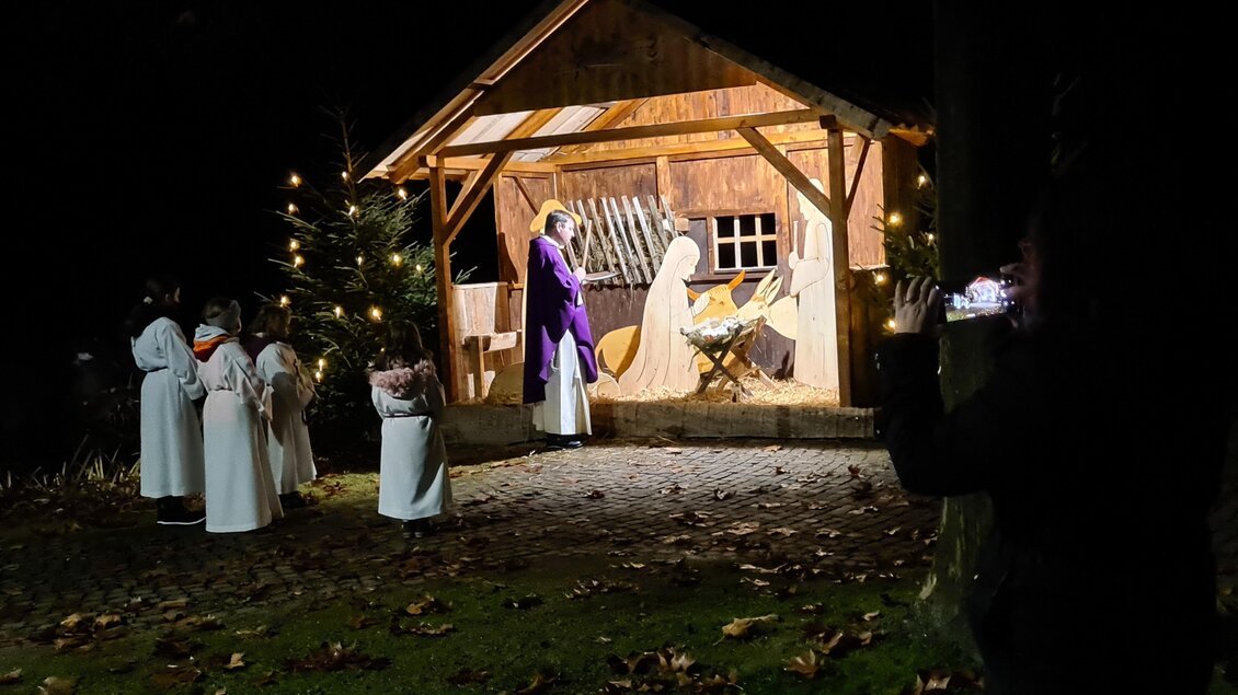 Eine stilisierte Krippe mit beleuchteter Hütte im Freien. Menschen in weißen Gewändern stehen in der Nähe und beobachten die Szene. | © Robier Johann| „Kripperlroas Oberhaag“